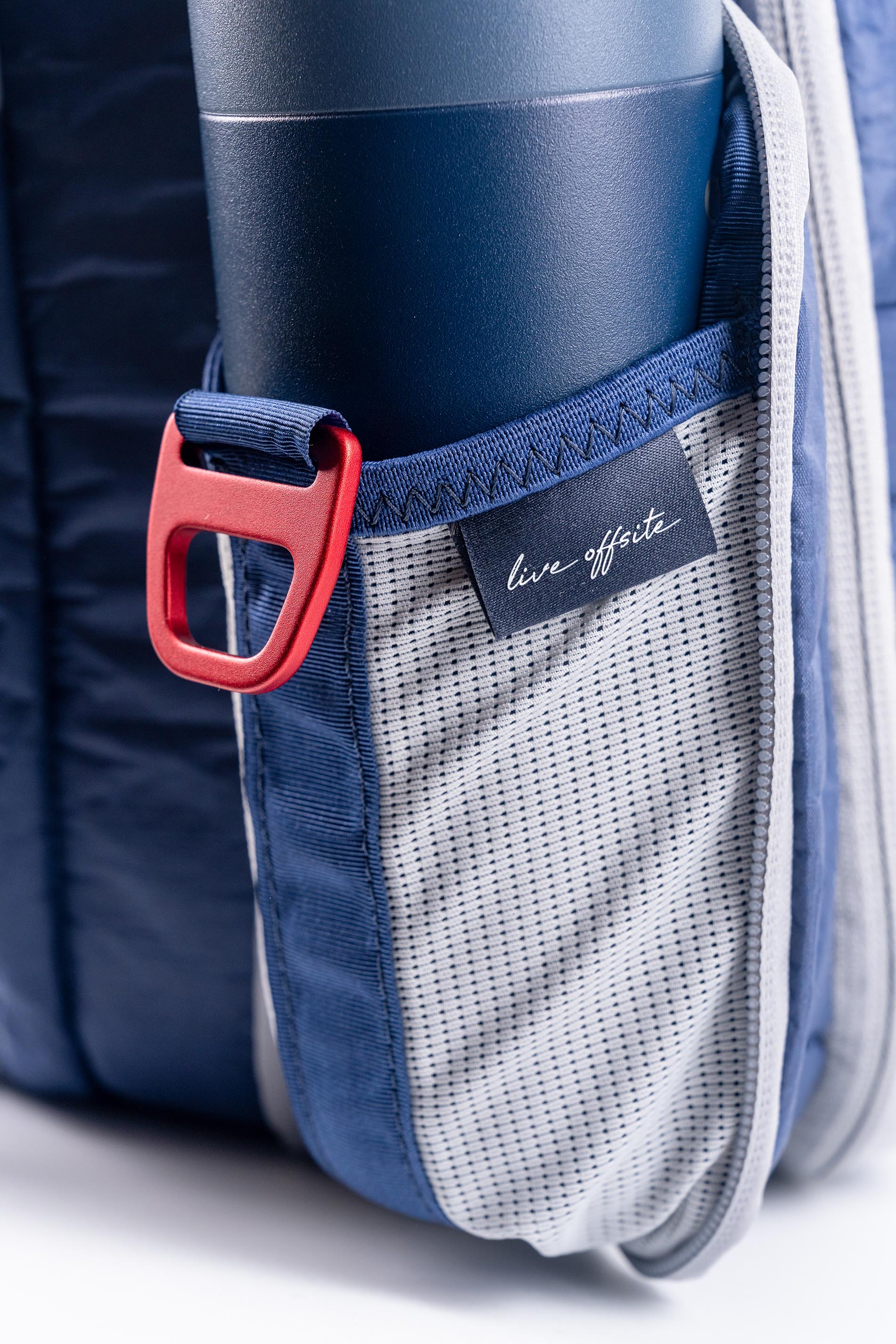 Close-up of a blue backpack with a red buckle and 'live offsite' label and pocket holding water bottle.